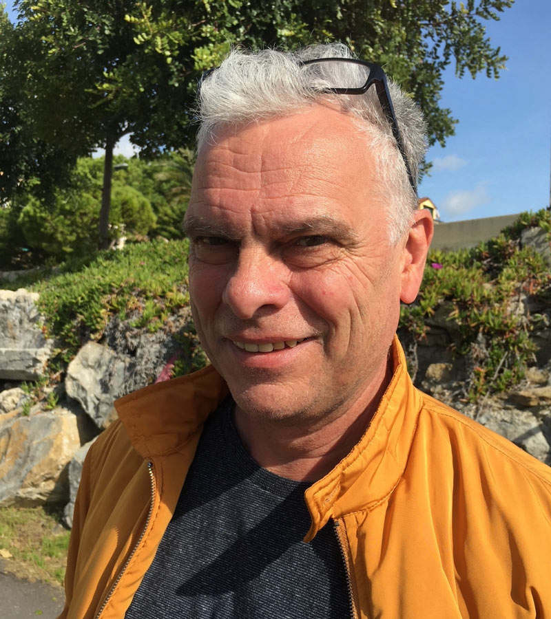Peter-Michael Peacock lächelnd im Freien vor einem bewachsenen Steinhang